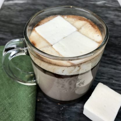 Hot chocolate with melted vanilla marshmallows in a clear glass mug setting on a dark marble board with a green napkin