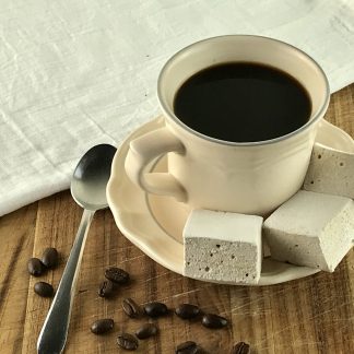 Three coffee marshmallows setting on a saucer with a cup of coffee. Coffee beans and a spoon are to the left of the saucer.