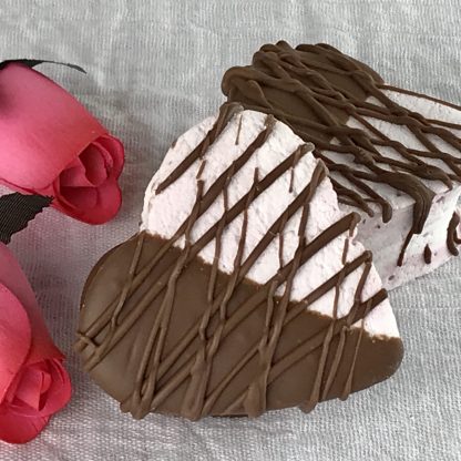 Two large marshmallow hearts half dipped in milk chocolate with additional drizzled chocolate setting on a white cloth with pink roses
