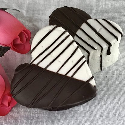 Two large marshmallow hearts half dipped in dark chocolate with additional drizzled chocolate setting on a white cloth with pink roses