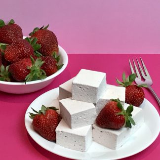Strawberry marshmallows and strawberries on a white plate setting on a pink background. There is also a white bowl of fresh strawberries.