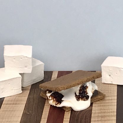 A s'more setting on a striped hardwood board. There vanilla marshmallows also setting on the board. The background is light gray.