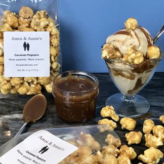 There is a dish of ice cream with caramel sauce drizzled on it and with a few pieces of caramel popcorn. There is also a small jar of caramel sauce with a spoon in it and an unopened bag of caramel popcorn. Lastly, there is an open bag of caramels popcorn with some spilled on the table.