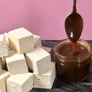 A stack of salted caramel marshmallows on a marble board with a pink background. There is a small jar of salted caramel sauce. A spoon has been dipped into the sauce and is held above the jar, allowing the sauce to drip into the jar.