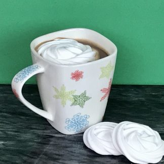 A mug of hot cocoa sets on a dark marble board. A white marshmallow flower sets in the hot cocoa. Two more marshmallow flowers set on the board. The background is green.