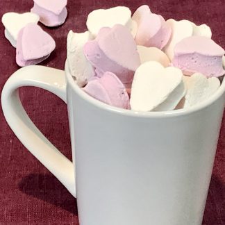A white mug is filled with mini marshmallow hearts in pink and white. The mug is setting on a burgundy linen. There are a few mini marshmallow hearts scattered on the linen cloth.