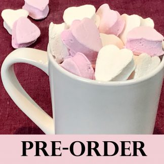A white mug is filled with mini marshmallow hearts in pink and white. The mug is setting on a burgundy linen napkin. There are a few mini marshmallow hearts scattered on the linen cloth. The word 'PRE-ORDER' is across the bottom of the photo.
