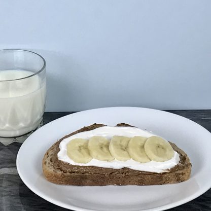 A slice of sourdough bread is slathered with almond butter and vanilla marshmallow whip. There are banana slices on top. The bread sets on a white plate, which is on a dark marble board. A glass of milk sets next to the plate.