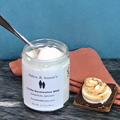 A spoon sets in an open jar of Vanilla Marshmallow Whip. Next to the jar is an open-face s'more consisting of a gluten-free graham cracker topped with melted Belgian chocolate and toasted marshmallow whip.
