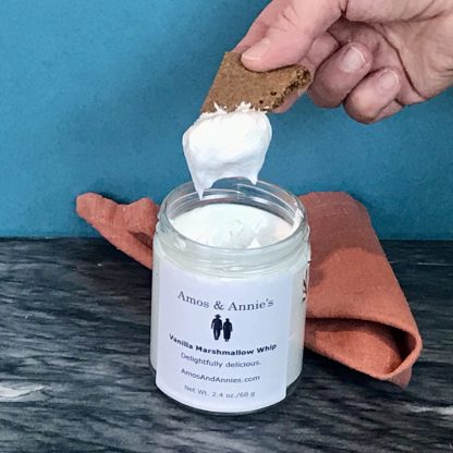 A hand holds a piece of gluten-free graham cracker which has been dipped into a jar of Vanilla Marshmallow Whip. The jar sets on a dark marble board with an orange napkin.