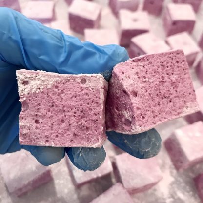 A gloved hand holds two wild blueberry marshmallows with other marshmallows in the background.