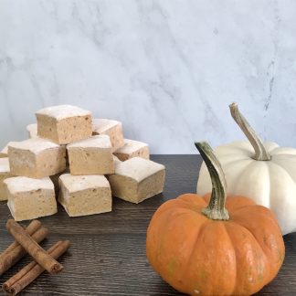 A pile of pumpkin spice marshmallows sets on a wooden board. Two miniature pumpkins and three cinnamon sticks are also on the board. The background is white marble.