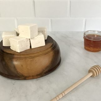 Wildflower honey marshmallows set on top of a upside-down wooden bowl. Next to the bowl is a small jar of honey and a wooden honey stirrer.