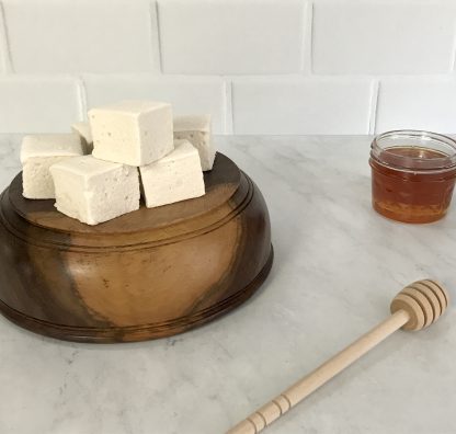 Wildflower honey marshmallows set on top of a upside-down wooden bowl. Next to the bowl is a small jar of honey and a wooden honey stirrer.