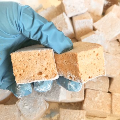 A gloved hand holds two pumpkin spice marshmallows with other marshmallows in the background.