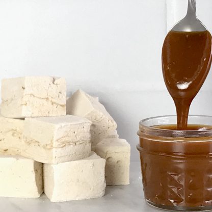 A stack of salted caramel marshmallows sets on a white marble surface with a white tile background. There is a small jar of salted caramel sauce. A spoon has been dipped into the sauce and is held above the jar, allowing the sauce to drip into the jar.