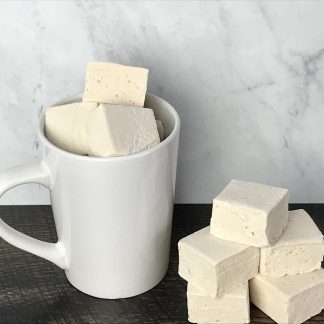 A white mug filled with peanut butter marshmallows sits on a dark board. Additional peanut butter marshmallows are stacked in the foreground near the mug. There is a light gray marble background.