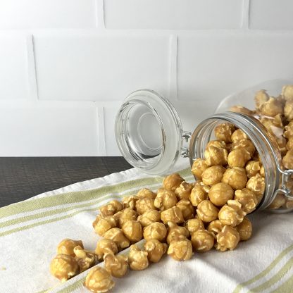A glass jar containing caramel popcorn is sitting on its side with the popcorn spilling out. The jar and popcorn are on a green and white striped towel, which is on a dark brown board. There is a white tile background.