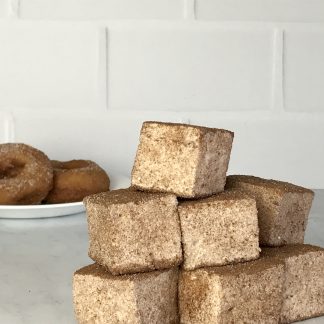A stack of apple cider donut marshmallows sits on a white marble board. There is a white plate with apple cider donuts in the background.