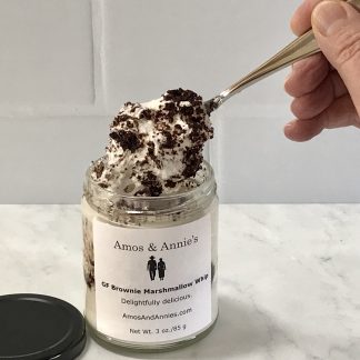 A jar of marshmallow whip with brownie pieces sits on a light gray marble board. A spoonful of brownie whip has been drawn out of the jar. There is a white tile background.