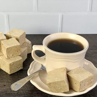 Two coffee marshmallows and a spoon sit on a saucer with a cup of coffee. A pile of coffee marshmallows are to the left of the saucer. All are on a brown board. There is a white subway tile background.
