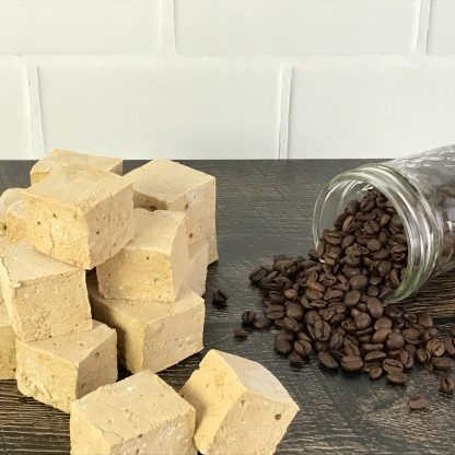 A pile of coffee marshmallows sits on a brown board with a jar of spilled coffee beans. The background is white subway tile.