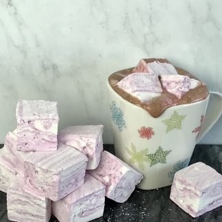 A mug of hot cocoa sits on a dark marble board. There are three peppermint marshmallows in the hot cocoa. The cocoa is just starting to drip over the edge of the mug. There are peppermint marshmallows stacked on the board. There is a light gray background.