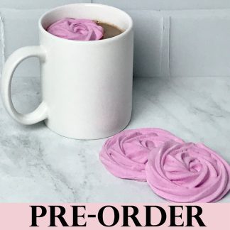 A mug of hot cocoa sits on a light gray marble board. A pink marshmallow flower floats in the hot cocoa. Two more marshmallow flowers are on the board. The background is white subway tile. The word 'PRE-ORDER' is across the bottom of the photo.