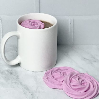 A mug of hot cocoa sits on a light gray marble board. A pink marshmallow flower floats in the hot cocoa. Two more marshmallow flowers are on the board. The background is white subway tile.