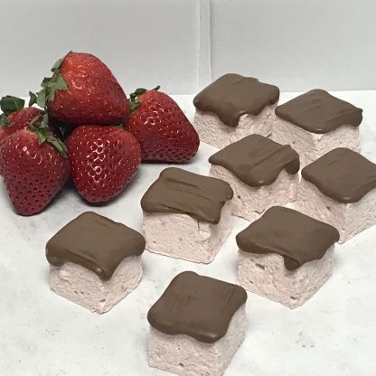 Strawberry marshmallows topped with Belgian milk chocolate sit on a white marble board. Fresh strawberries are piled next to the marshmallows. There is a white tile background.