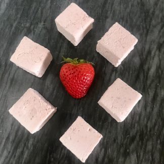 Six strawberry marshmallows are arranged in a circle on a dark marble board. A fresh strawberry is in the middle of the circle.
