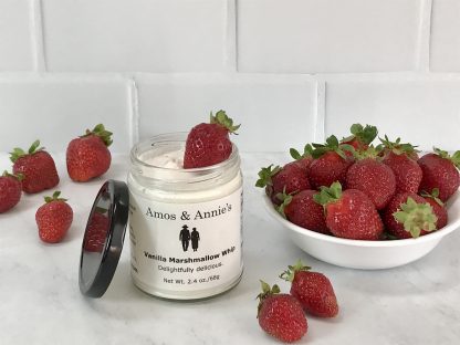 An opened jar of vanilla marshmallow whip sits on a light gray marble board. The jar’s black lid is resting against the jar. A small bowl of strawberries is to the right of the jar. There are several strawberries scattered around the board. There is a white tile background.