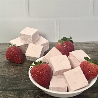 A white bowl of strawberry marshmallows and fresh strawberries sits on a dark wooden board. Additional strawberry marshmallows are piled on the board with a fresh berry next to them. There is a white tile background.