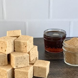 A stack of maple marshmallows sits on a brown board. There is a small jar of maple syrup and another of maple sugar. The marshmallows have a maple sugar coating on all sides. The background is white subway tile.