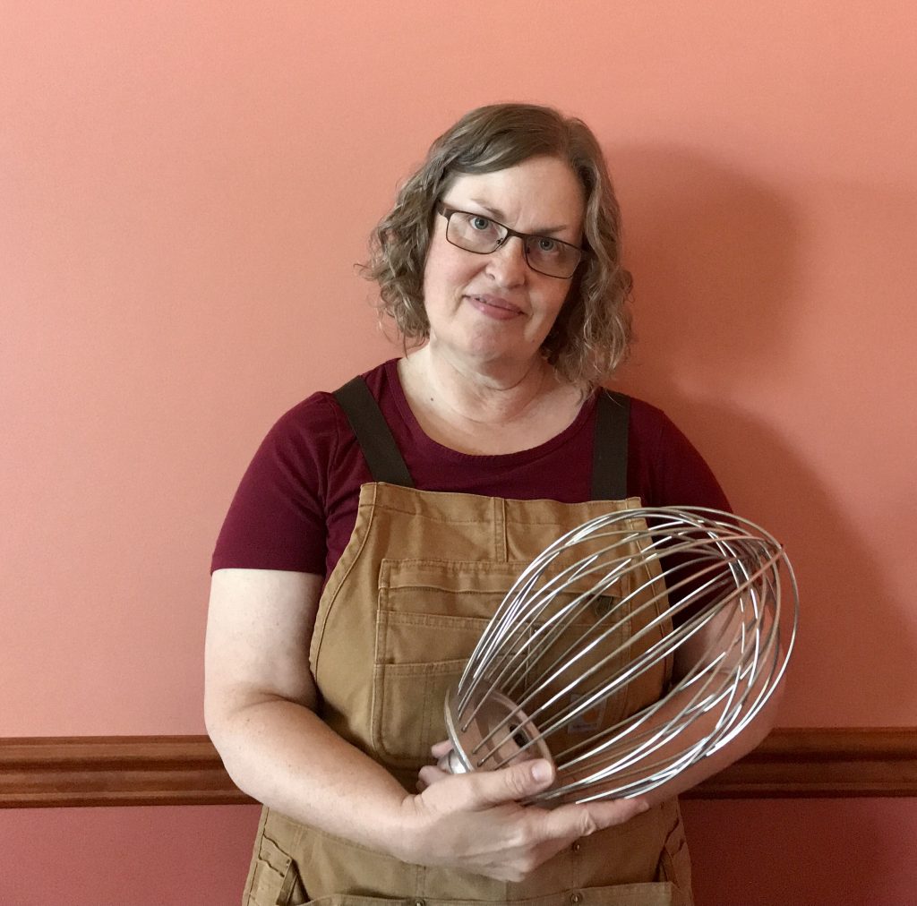 Amos and Annie’s owner Lisa is wearing an apron and holding a large whisk for a commercial mixer. The background is a painted wall with chair rail.