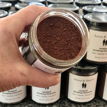 A hand is holding a jar of hot cocoa mix. The jar is opened showing the contents. There are bits of chocolate in the mix. Additional jars of hot cocoa mix are in the background.