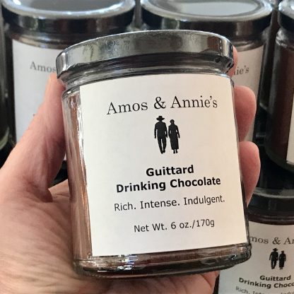 A hand is holding a jar of hot cocoa mix. The jar is clear and has a black lid. Additional jars of hot cocoa mix are in the background.