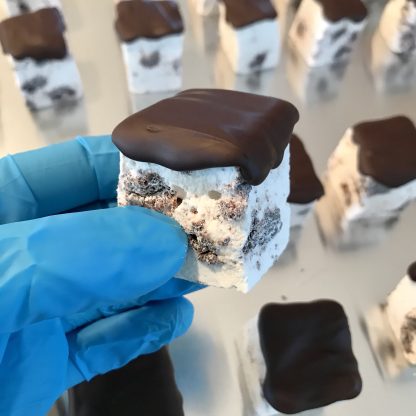A gloved hand is holding a ChocoMint cookie marshmallow. Additional ChocoMint cookie marshmallows are lined up on a baking sheet in the background. The marshmallows contain gluten-free cookie pieces and are topped with Belgian dark chocolate.
