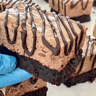 A gloved hand holds a marshmallow brownie bar. The bottom layer is a gluten-free brownie. The top layer is chocolate marshmallow. There is a Belgian dark chocolate drizzle over the marshmallow.