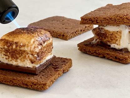Toasted vanilla marshmallows sit atop homemade graham crackers with chocolate. A flame from a kitchen torch is toasting one of the marshmallows.