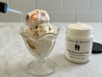 There is a clear glass bowl with ice cream topped with vanilla marshmallow whip. A small kitchen torch is toasting the whip. There is also a full jar of marshmallow whip to the side. The surface is gray marble. The background is white subway tile.