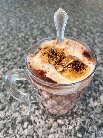 Hot cocoa is in a clear, glass mug. There are toasted vanilla marshmallows on top. A spoon is also in the mug.