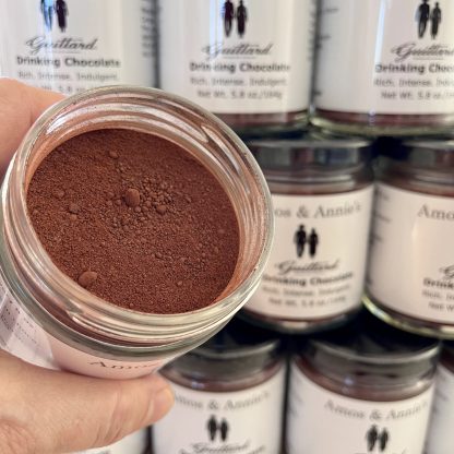 A hand is holding a jar of hot cocoa mix. The jar is opened showing the contents. There are bits of chocolate in the mix. Additional jars of hot cocoa mix are in the background.