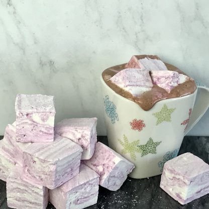 Hot cocoa is in a white ceramic mug. There are peppermint marshmallows on top. There is a pile of the marshmallows next to the mug. The background is gray marble.