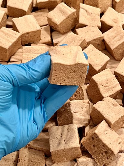 A gloved hand is holding a gingerbread marshmallow. There is a pile of gingerbread marshmallows in the background.