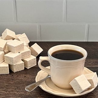 Two coffee marshmallows and a spoon sit on a saucer with a cup of coffee. A pile of coffee marshmallows are to the left of the saucer. All are on a brown board. There is a white subway tile background.