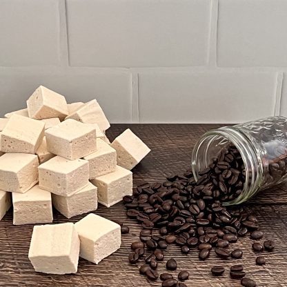 A pile of coffee marshmallows sits on a brown board with a jar of spilled coffee beans. The background is white subway tile.