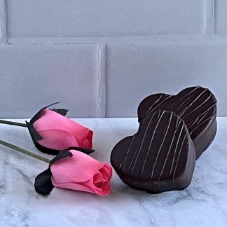 Two large marshmallow hearts dipped in dark chocolate sit on light gray marble with artificial pink roses nearby.