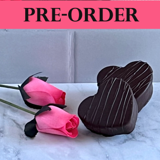 Two large marshmallow hearts dipped in dark chocolate sit on light gray marble with artificial pink roses nearby. The word ‘pre-order’ is at the top of the photo.