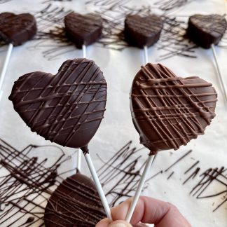 A hand holds two marshmallow heart lollipops. The marshmallow hearts are coated in Belgian chocolate. The background is a pan of more lollipops.